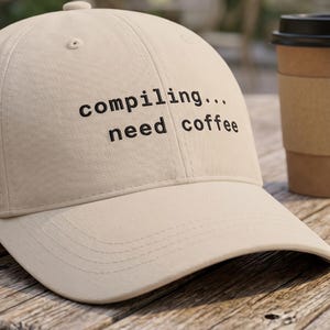 Puede incluir: Gorra de béisbol beige con el texto "compiling... need coffee" en negro. Una taza de café desechable con una funda marrón y tapa negra está al lado de la gorra. Sobre una mesa de madera desgastada.