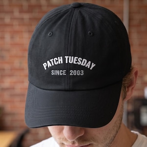 May include: A black baseball cap featuring the white embroidered text PATCH TUESDAY arched over the smaller text SINCE 2003. The cap has a structured crown, a curved brim, and a button at the top center, providing a clean and simple design.