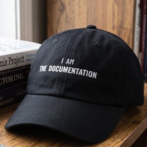 May include: A black baseball cap featuring the white embroidered text I AM THE DOCUMENTATION on the front panel. The cap has a structured crown, a curved brim, and a button on top, presented in a clean, minimalist style for everyday wear.