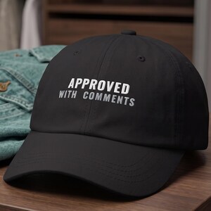 May include: A black baseball cap featuring the white text "APPROVED WITH COMMENTS" embroidered on the front panel. The cap has a curved brim and a structured crown, sitting on a wooden surface next to a folded blue denim jacket.