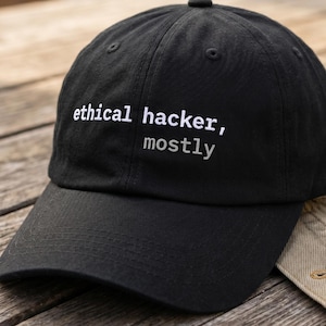 May include: Black baseball cap with the text "ethical hacker, mostly" embroidered in white. The cap is resting on a wooden surface next to a tan jacket.