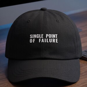 May include: A black baseball cap featuring the white embroidered text "SINGLE POINT OF FAILURE" centred on the front panel. The cap has a structured crown, a curved brim, and a button at the top, constructed from a solid black fabric material.