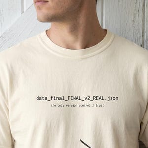 May include: A cream-coloured short-sleeve t-shirt featuring the black printed text data_final_FINAL_v2_REAL.json and the smaller text the only version control i trust centred on the chest area, with a crew neckline and a standard fit.