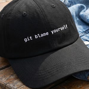 May include: A black baseball cap with the white embroidered text "git blame yourself". The cap is on a wooden surface next to a denim jacket. The cap has a curved peak and a button on top.
