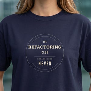 Peut inclure: T-shirt bleu marine avec un motif circulaire. Le motif comprend le texte "THE REFACTORING CLUB", "ESTABLISHED" et "NEVER". Le t-shirt est de couleur unie et a des manches courtes.