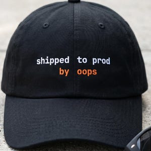 Può includere: Cappellino da baseball nero con la scritta "shipped to prod by oops" in bianco e arancione. Il cappellino è appoggiato su una superficie di cemento accanto a un paio di occhiali da sole neri.