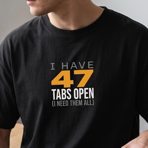 May include: A black short-sleeved t-shirt featuring the text "I HAVE 47 TABS OPEN (I NEED THEM ALL)" printed on the chest. The words "I HAVE" and "(I NEED THEM ALL)" are in white, while the number "47" is in orange and "TABS OPEN" is in white.