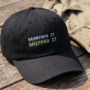 May include: A black baseball cap with the text "SEARCHED IT SHIPPED IT" embroidered in white and green. The cap is sitting on a wooden surface next to a tan denim jacket.