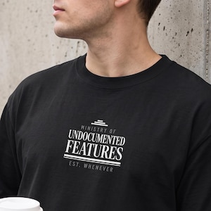 May include: A black short-sleeved t-shirt featuring white text on the chest that reads Ministry of Undocumented Features Est. Whenever. The design includes horizontal lines above and below the main text, creating a clean, centered graphic.