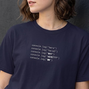 May include: A dark navy blue short-sleeved t-shirt featuring white printed code text on the chest that reads: console.log("here"), console.log("here2"), console.log("WHY"), console.log("HERE???"), and console.log("👀"). The shirt has a crew neckline.