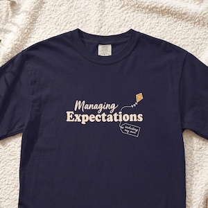 May include: A navy blue short-sleeve t-shirt featuring the text "Managing Expectations" in a light cream font. A small graphic of a kite and a rectangular tag with the text "including my own" are printed in white next to the main text.