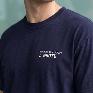 Può includere: T-shirt blu navy con la scritta bianca "REPLACED BY A SCRIPT I WROTE". La maglietta ha un girocollo e maniche corte. Il tessuto sembra essere un morbido misto cotone.