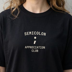 Könnte beinhalten: Schwarzes T-Shirt mit dem weißen Text "SEMICOLON APPRECIATION CLUB". Das Design beinhaltet ein Semikolon-Symbol. Das Shirt ist ein lässiger Stil mit kurzen Ärmeln.
