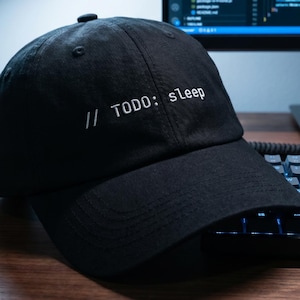 Peut inclure: Casquette de baseball noire avec l'inscription blanche "// TODO: sleep". La casquette est posée sur un bureau en bois, à côté d'un clavier et d'un écran d'ordinateur.