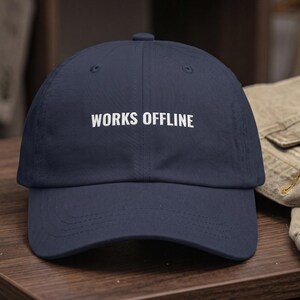 May include: A navy blue baseball cap featuring the white embroidered text WORKS OFFLINE centered on the front panel. The cap has a structured crown, a curved brim with visible stitching, and a button at the top center of the dome.
