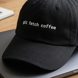 Puede incluir: Gorra de béisbol negra con el texto "git fetch coffee" en blanco. La gorra está sobre un portátil en una superficie de madera. Un cuaderno y una taza también son visibles en el fondo.