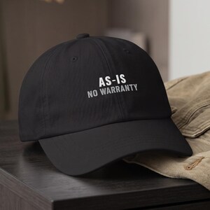 May include: A black baseball cap with a curved brim and a structured crown. The front of the cap features the text "AS-IS" in white capital letters, with the words "NO WARRANTY" printed directly underneath in a smaller white font.