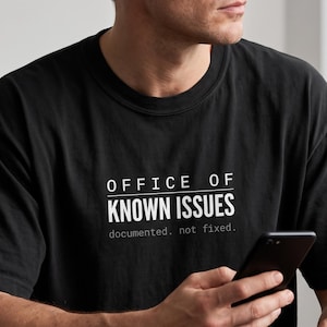 May include: A black short-sleeved t-shirt featuring the white text "OFFICE OF KNOWN ISSUES" in a bold, sans-serif font, with the smaller text "documented. not fixed." printed directly underneath in a lighter, typewriter-style font.