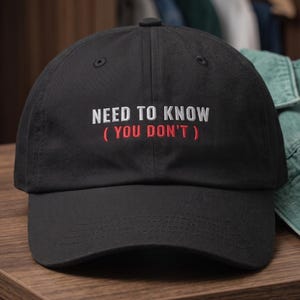 May include: A black baseball cap featuring embroidered text on the front panel. The top line reads NEED TO KNOW in white capital letters, and the bottom line reads ( YOU DON'T ) in red capital letters. The cap has a structured crown and a curved brim.