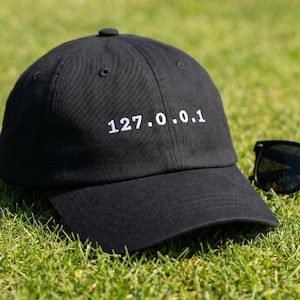 May include: Black baseball cap with the text "127.0.0.1" in white. The cap is on a bed of green grass next to a pair of black sunglasses.