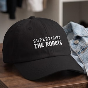 May include: A black baseball cap featuring the white embroidered text SUPERVISING THE ROBOTS on the front panel. The cap has a curved brim, a structured crown with eyelets, and a button at the top, presented in a classic six-panel design.