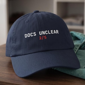 Peut inclure: Une casquette de baseball bleu marine avec le texte brodé en blanc "DOCS UNCLEAR" et le texte brodé en rouge "2/5" juste en dessous. La casquette présente une calotte structurée, une visière incurvée et des détails de couture visibles.