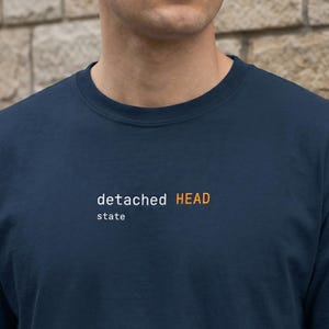 May include: A navy blue t-shirt featuring the text "detached HEAD state" in white and orange. The shirt has a crew neck and is made of a soft fabric.