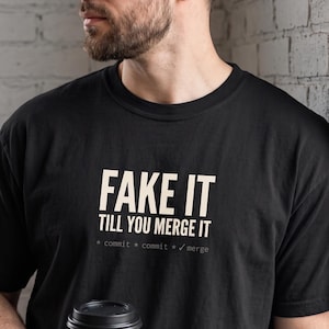 Puede incluir: Una camiseta negra de manga corta con el texto en color beige "FAKE IT TILL YOU MERGE IT" impreso en el pecho, seguido del texto más pequeño "* commit * commit * ✓ merge" en una fuente estilo máquina de escribir, centrado en la parte frontal.