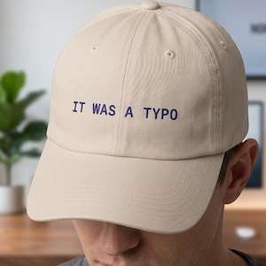 Op de afbeelding: Een beige baseballcap met de geborduurde tekst "IT WAS A TYPO" in marineblauw. De cap heeft een gebogen klep en een knoop aan de bovenkant. De cap wordt door een persoon gedragen.