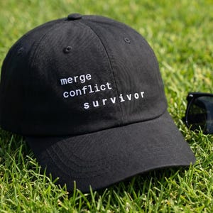 May include: A black baseball cap with the words "merge conflict survivor" embroidered in white. The cap is on green grass, next to a pair of black sunglasses.