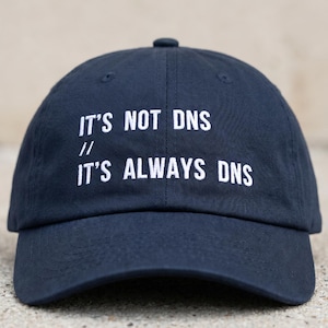 Puede incluir: Una gorra de béisbol azul marino con texto bordado en blanco en la parte frontal que dice IT'S NOT DNS // IT'S ALWAYS DNS. La gorra tiene una corona estructurada, una visera curva y un botón en la parte superior, hecha de tela sólida.