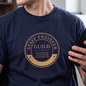 May include: A navy blue t-shirt featuring a circular gold emblem with the text STAFF ENGINEER GUILD. Below the text, it reads: Writes documents. Attends meetings. Occasionally codes. The design includes decorative lines and small star accents.