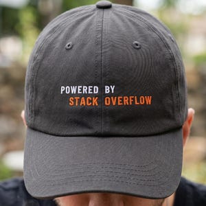 May include: A charcoal grey baseball cap featuring the text "POWERED BY STACK OVERFLOW" in white and orange. The cap is worn by a person holding a mug.