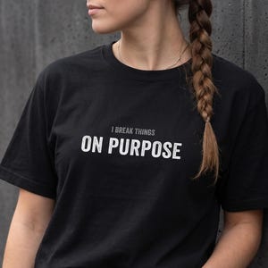 May include: A black short-sleeved t-shirt featuring the text I BREAK THINGS ON PURPOSE printed in white and grey on the chest. The shirt is paired with blue denim jeans that have a visible metal button and rivets at the waistline.
