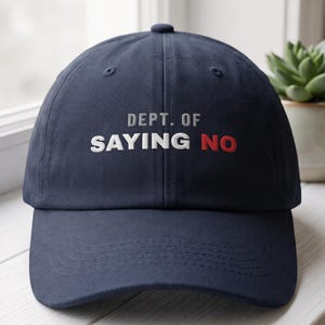 May include: A navy blue baseball cap featuring the embroidered text DEPT. OF SAYING NO on the front. The word DEPT. OF is in grey, SAYING is in white, and NO is in red. The cap has a structured crown, a curved brim, and a button on top.