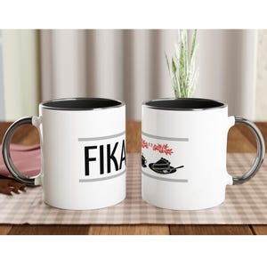 Könnte beinhalten: Zwei weiße Keramikbecher mit schwarzem Innenraum und silberfarbenen Griffen stehen auf einem Holztisch. Auf einem Becher steht das Wort "FIKA" in schwarzen Buchstaben. Der andere Becher hat eine Grafik einer Kaffeetasse und Gebäck.