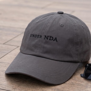 May include: A charcoal gray baseball cap featuring the text "UNDER NDA" embroidered in black thread on the front panel. The cap has a structured crown, a curved brim, and a button at the top. Black sunglasses are positioned next to the cap.