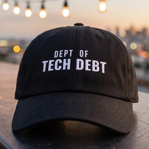 Op de afbeelding: Een zwarte baseballpet met de wit geborduurde tekst DEPT OF TECH DEBT in het midden op het voorpaneel. De pet heeft een gestructureerde kroon, een gebogen klep en zichtbare stiksels op de stoffen panelen en de klep.