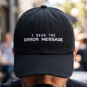 May include: A black baseball cap featuring the white embroidered text I READ THE ERROR MESSAGE centered on the front panel. The cap has a structured crown, a curved brim, and a button at the top, constructed from a solid black fabric material.