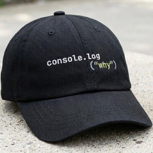 Może przedstawiać: Czarna czapka z daszkiem z napisem "console.log ("why")" w kolorze białym i jasnoniebieskim. Czapka leży na betonowej powierzchni obok kubka z kawą.