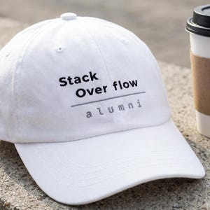 Könnte beinhalten: Eine weiße Baseballkappe mit dem Text "Stack Overflow alumni" in Schwarz. Neben der Kappe steht ein Einweg-Kaffeebecher mit brauner Hülle. Der Hut ist aus weichem Material.