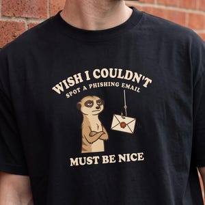Puede incluir: Una camiseta negra de manga corta con un gráfico de un suricato con los brazos cruzados mirando un sobre colgado de un anzuelo. El texto dice WISH I COULDN'T SPOT A PHISHING EMAIL MUST BE NICE en un color crema claro.