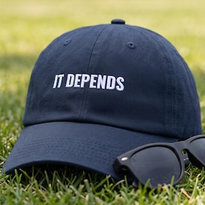 May include: A navy blue baseball cap featuring the white embroidered text IT DEPENDS on the front panel, accompanied by a pair of black sunglasses resting on the brim. The cap has a structured crown, a curved visor, and a button on the top.