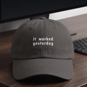 May include: A charcoal gray baseball cap featuring the white embroidered text "it worked yesterday" centered on the front panel. The cap has a structured crown, a curved brim, and stitched eyelets for ventilation on the top panels.