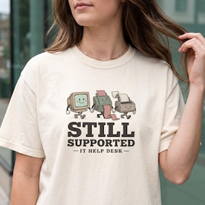 Peut inclure: Un t-shirt à manches courtes de couleur crème avec un motif représentant trois machines de bureau vintage avec des visages et des membres qui marchent ensemble. Sous le motif, le texte indique STILL SUPPORTED IT HELP DESK en gras.