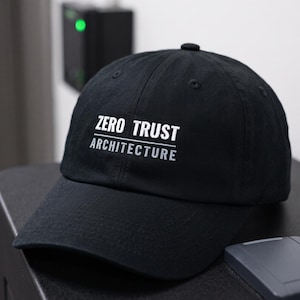 May include: A black baseball cap featuring the white text ZERO TRUST and the grey text ARCHITECTURE embroidered on the front. The cap has a structured crown, a curved brim, and a button on top, presenting a clean and professional appearance.