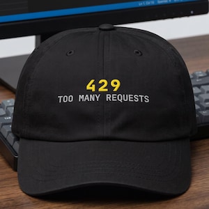 Pode incluir: Um boné de beisebol preto com o texto 429 bordado em amarelo e o texto TOO MANY REQUESTS bordado em branco no centro do painel frontal. O boné possui uma aba curva e uma copa estruturada com um botão no topo central.