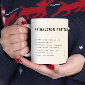 Puede incluir: Una taza de cerámica blanca con el texto "EXTRACTION PROTOCOL" y especificaciones técnicas. La taza es sostenida por una persona que lleva un suéter azul oscuro con un patrón de renos rojos.