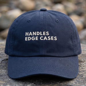May include: A navy blue baseball cap featuring the embroidered white text "HANDLES EDGE CASES" on the front panel. The cap has a structured crown, a curved brim, and a button at the top center, constructed from a durable fabric with visible stitching.
