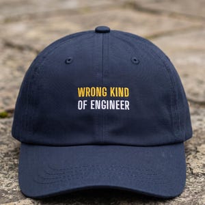 May include: A navy blue baseball cap featuring the embroidered text WRONG KIND in yellow and OF ENGINEER in white on the front panel. The cap has a structured crown, a curved brim, and a button at the top centre of the six-panel construction.